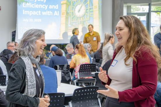 Two people conversing in a room full of people sitting down, where a presentation is about to take place